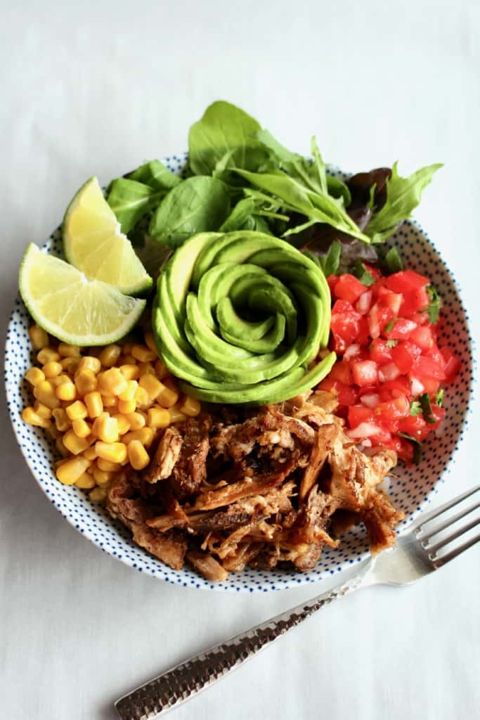 A plate with crispy carnitas, pico de gallo, corn, salad, avocado, and lime wedges.