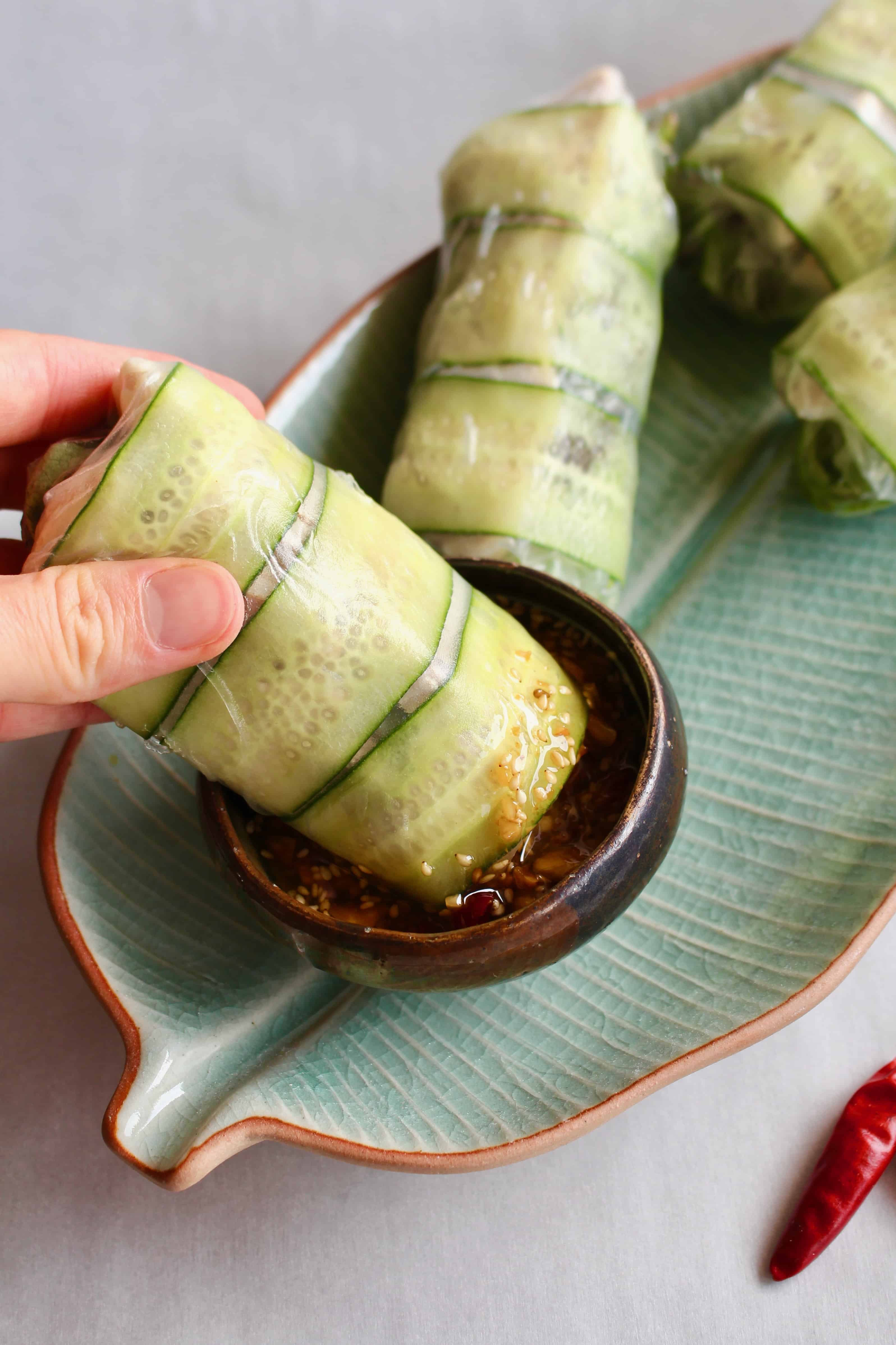 A hand dipping a summer roll with cucumbers into a dish of spicy sauce.