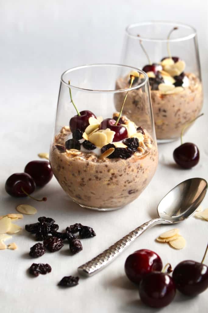 Glass tumblers with overnight oats inside with fresh cherries, dried cherries, and sliced almonds scattered around.