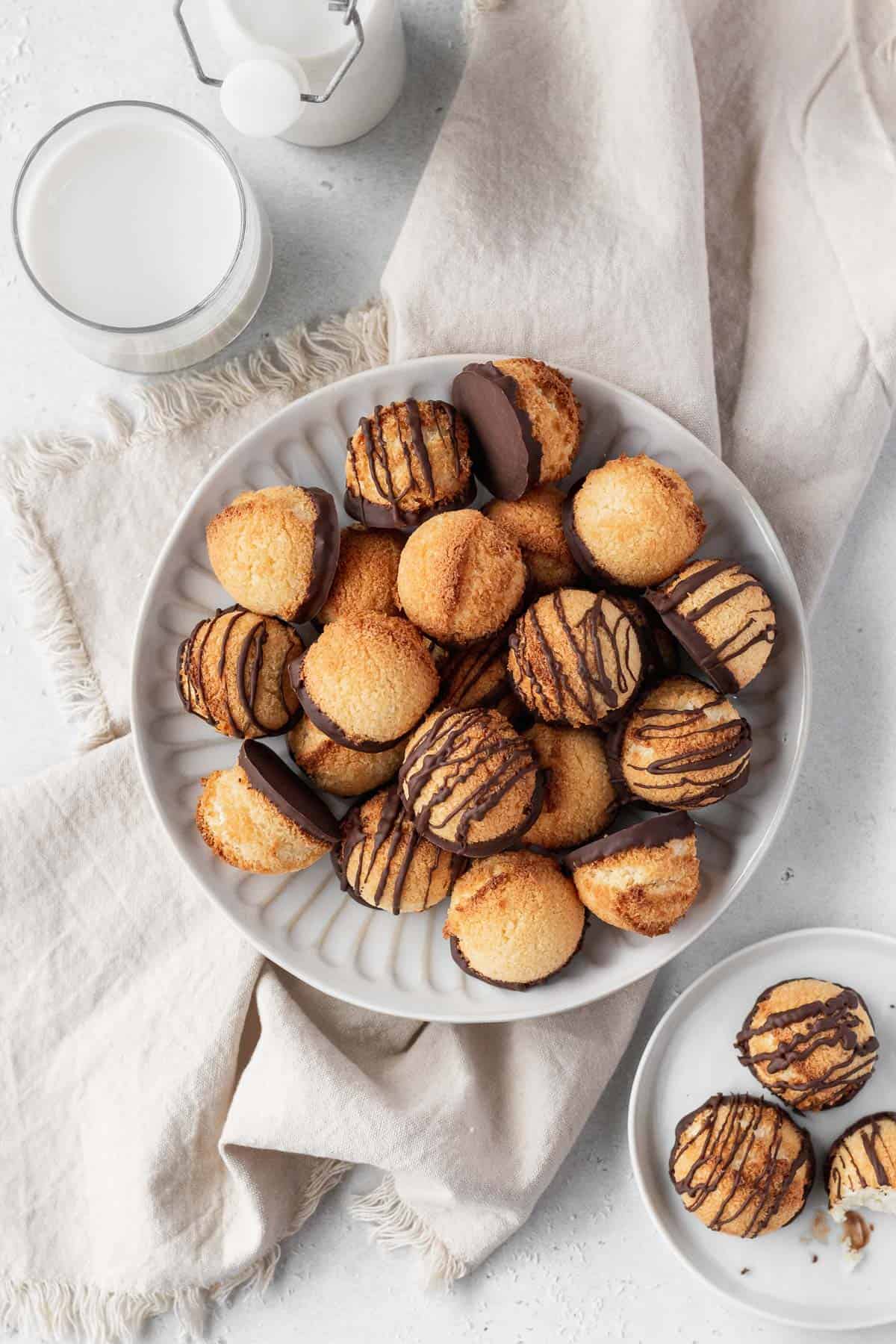 Overhead shot of plain and chocolate drizzled coconut macaroons on a plate with a glass of milk, a smaller plate of macaroons, and a natural linen.