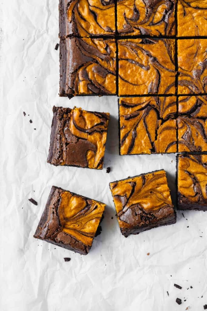 Three pieces of pumpkin cheesecake brownies scattered next to more brownies on a piece of parchment paper.