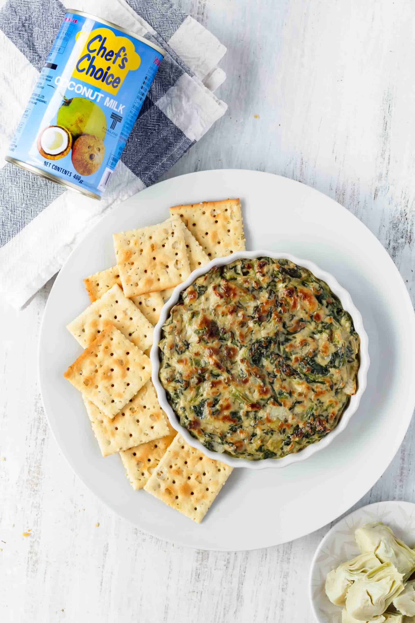 A dish of vegan spinach artichoke dip next to some crackers on a white plate, and a blue can of coconut milk laying above in the corner of the shot.
