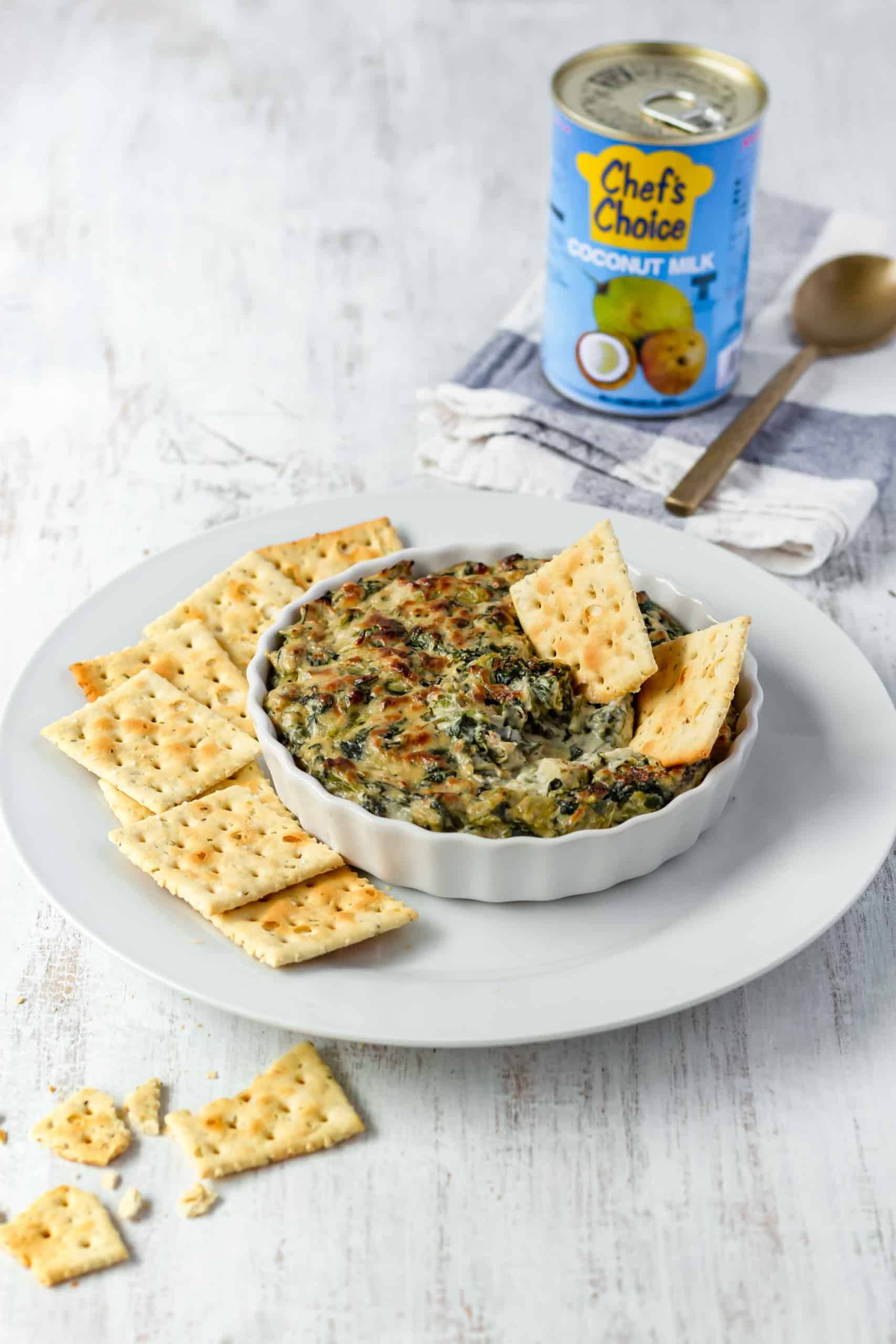 A plate of crackers with a white dish of hot spinach dip, with a can of coconut milk in the background.