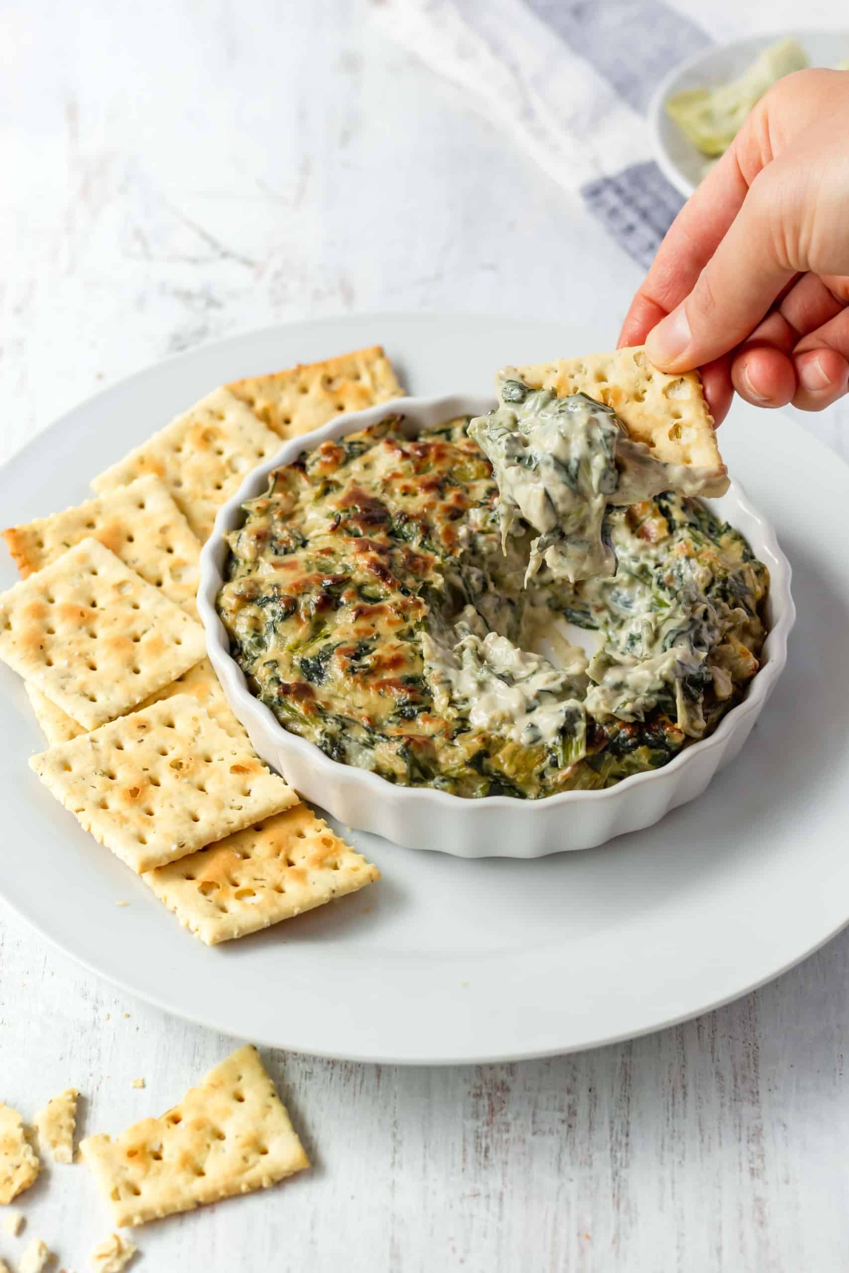 A hand holding a saltine cracker and dipping it into vegan spinach artichoke dip.