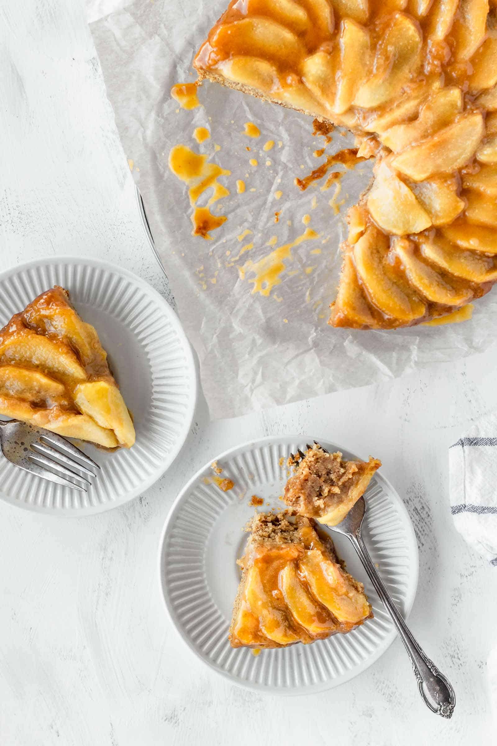 Two slices of vegan apple cake on small white plates next to the whole cake.