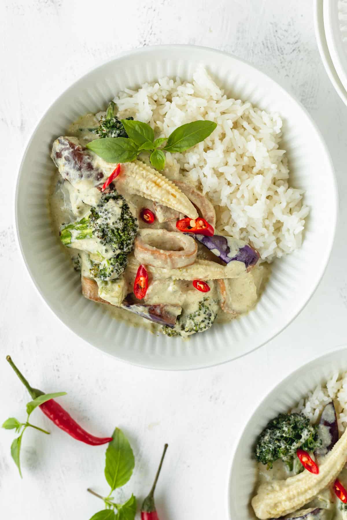 A white bowl of vegan Thai curry with rice on a white surface with some herbs and chili peppers scattered around.