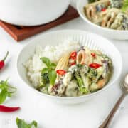 Square image of vegan Thai green curry in a white bowl with a silver spoon next to it.
