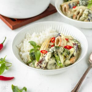 Square image of vegan Thai green curry in a white bowl with a silver spoon next to it.
