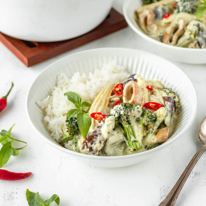 Square image of vegan Thai green curry in a white bowl with a silver spoon next to it.