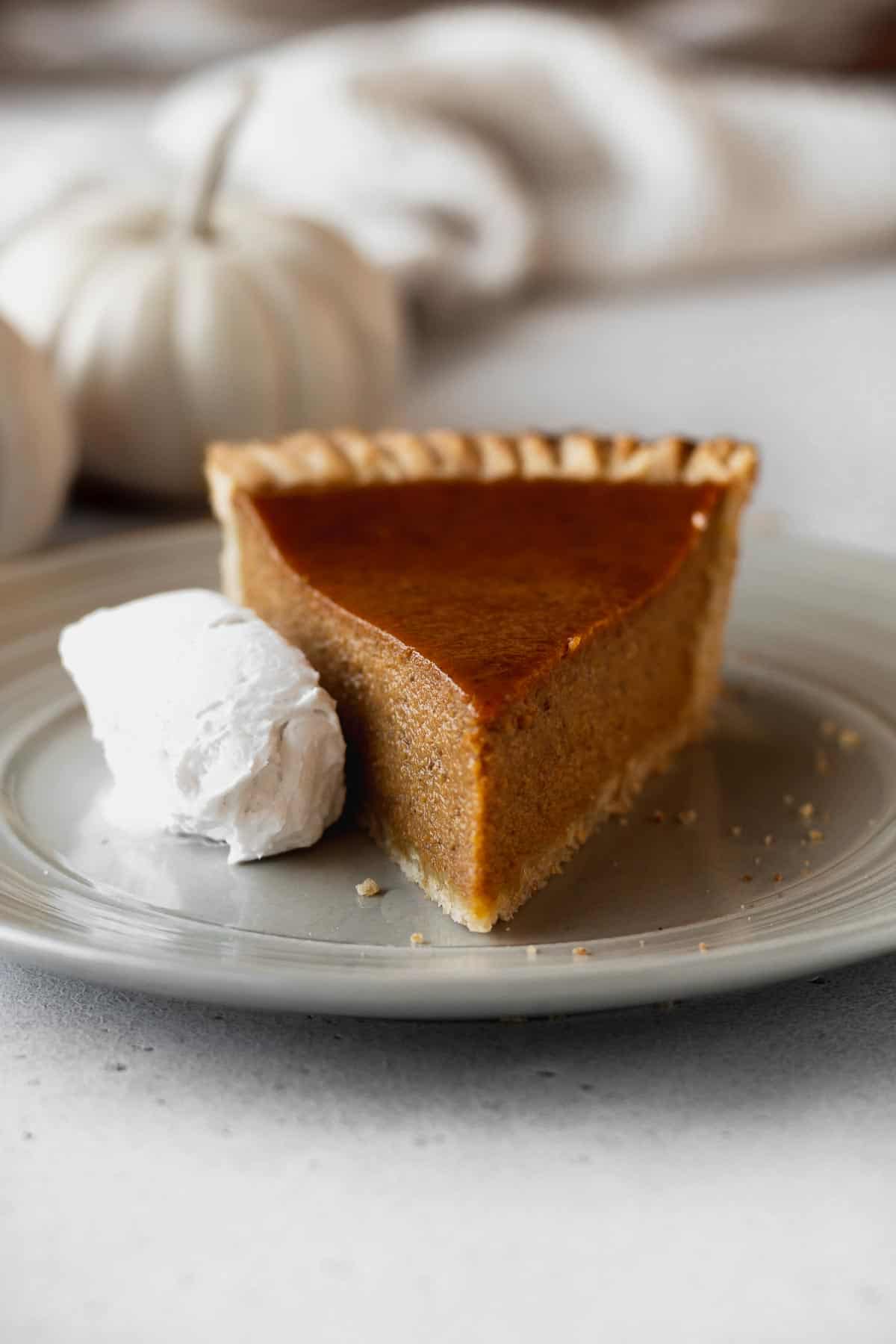 A slice of dairy-free pumpkin pie on a grey plate with a whipped coconut cream on the side and a white pumpkin in the background.