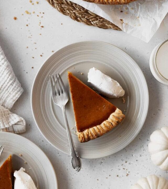 Overhead shot of a slice of dairy-free pumpkin pie with whipped cream and a fork on a plate surrounded by plates and white pumpkins.
