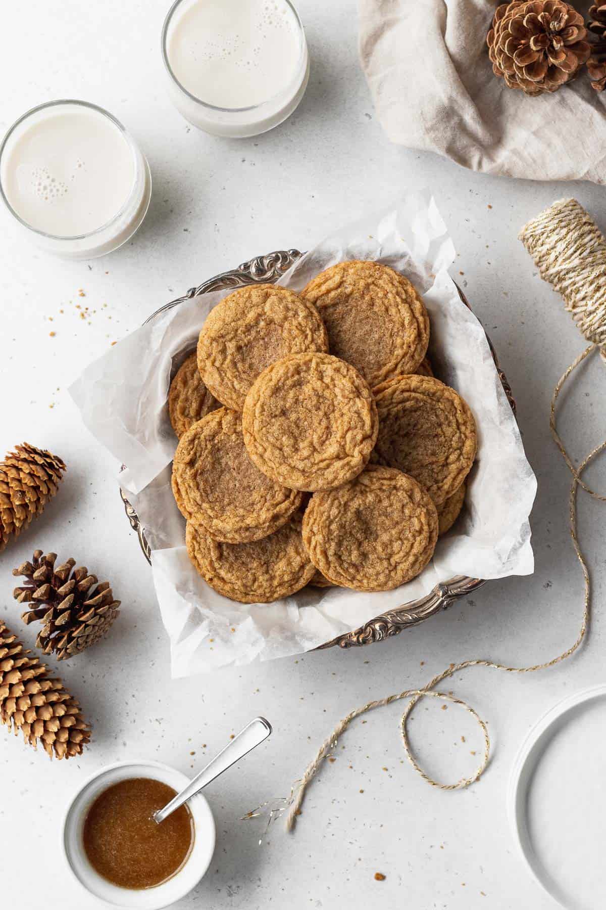 A platter of gluten free ginger snaps surrounded by glasses of milk, pine cones, string, and a dish of maple glaze icing.