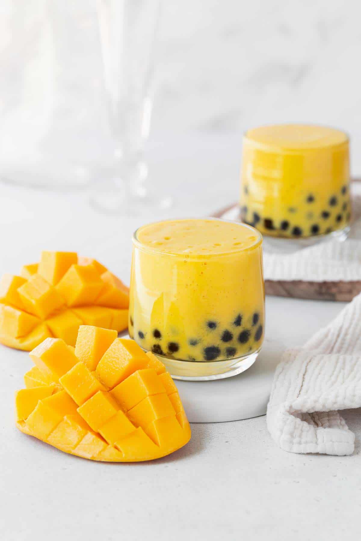 Two glasses of mango boba tea on a white counter next to a cubed up mango.