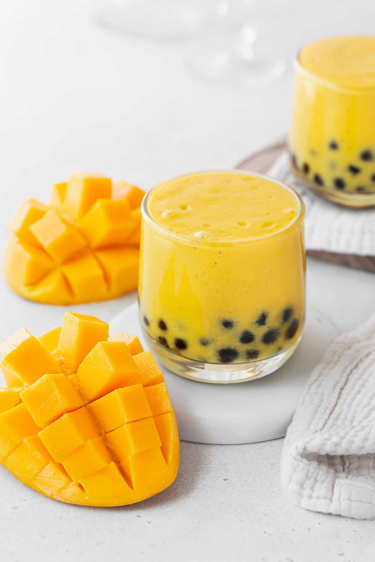 A closeup shot of a small glass of mango boba tea on a white coaster on the counter next to a sliced up mango.