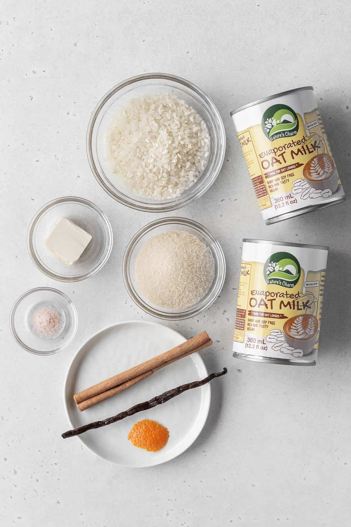 Vegan rice pudding ingredients measured out into bowls with 2 cans of evaporated oat milk on a white surface.