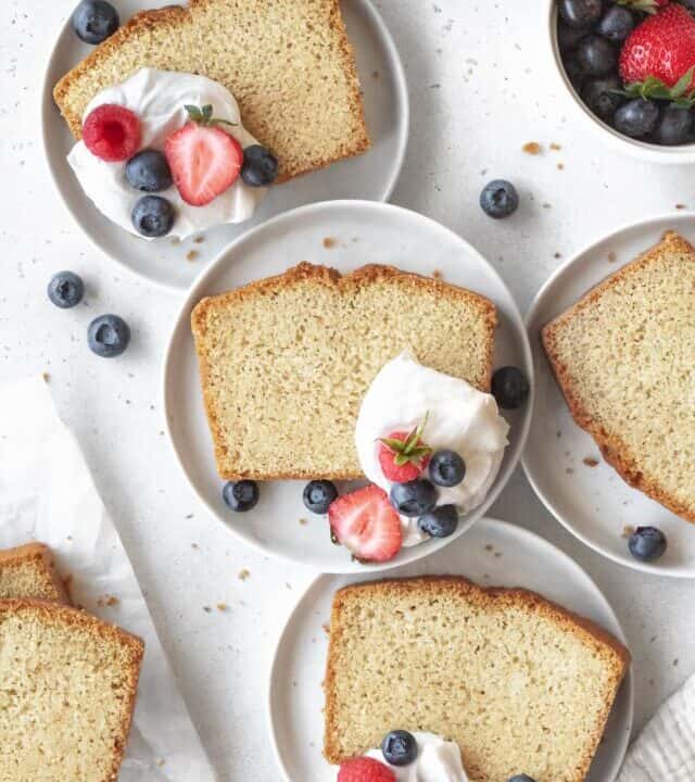 Slices of gluten free pound cake on small white plates with coconut cream and berries.