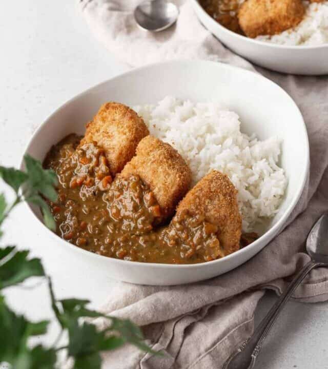 Vegan katsu curry in a white bowl on a linen with a flat leaf parsley plant in the corner of the shot.