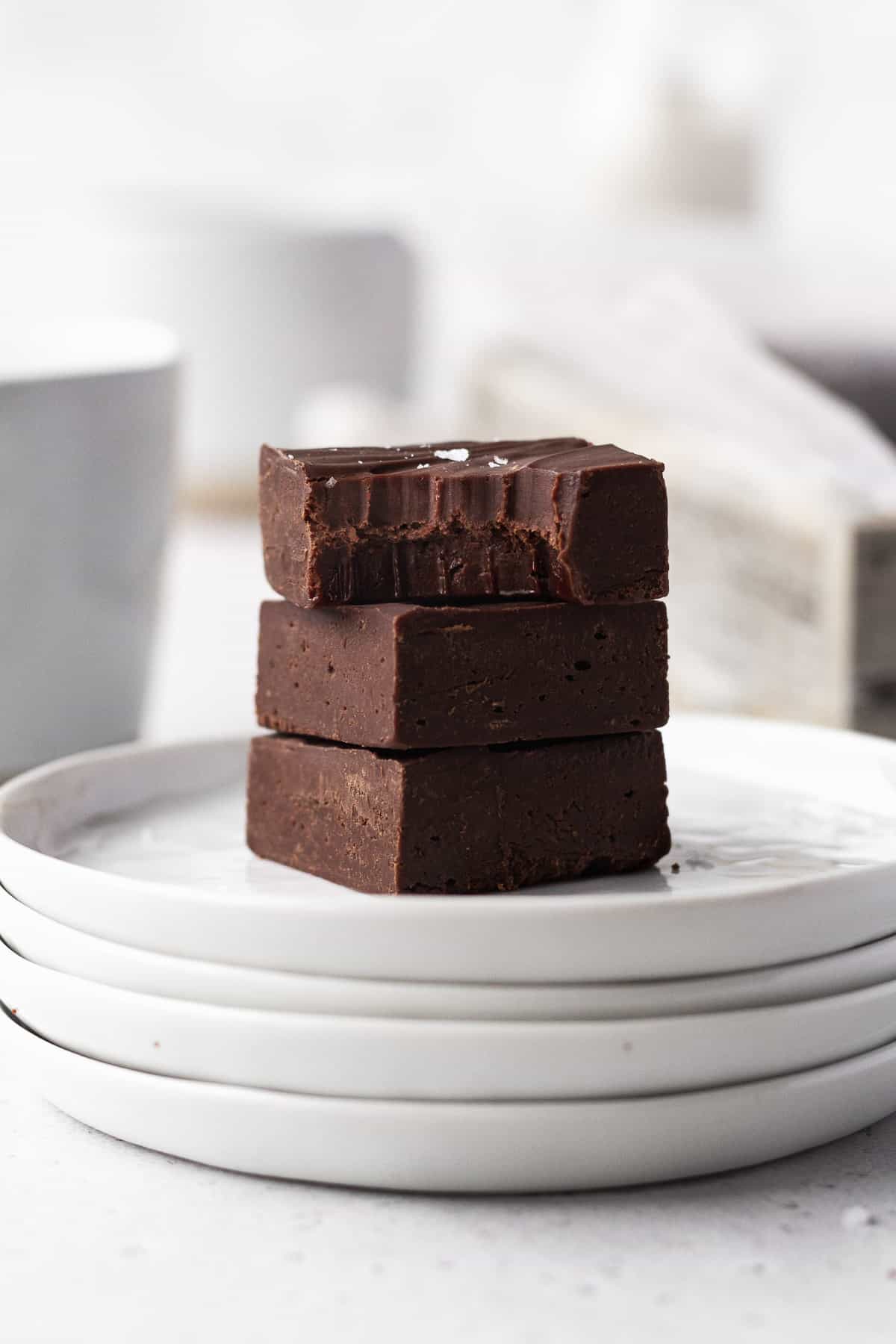 Side on shot of 3 cubes of dairy-free fudge stacked on a stack of white plates with the top square having a bite taken out.