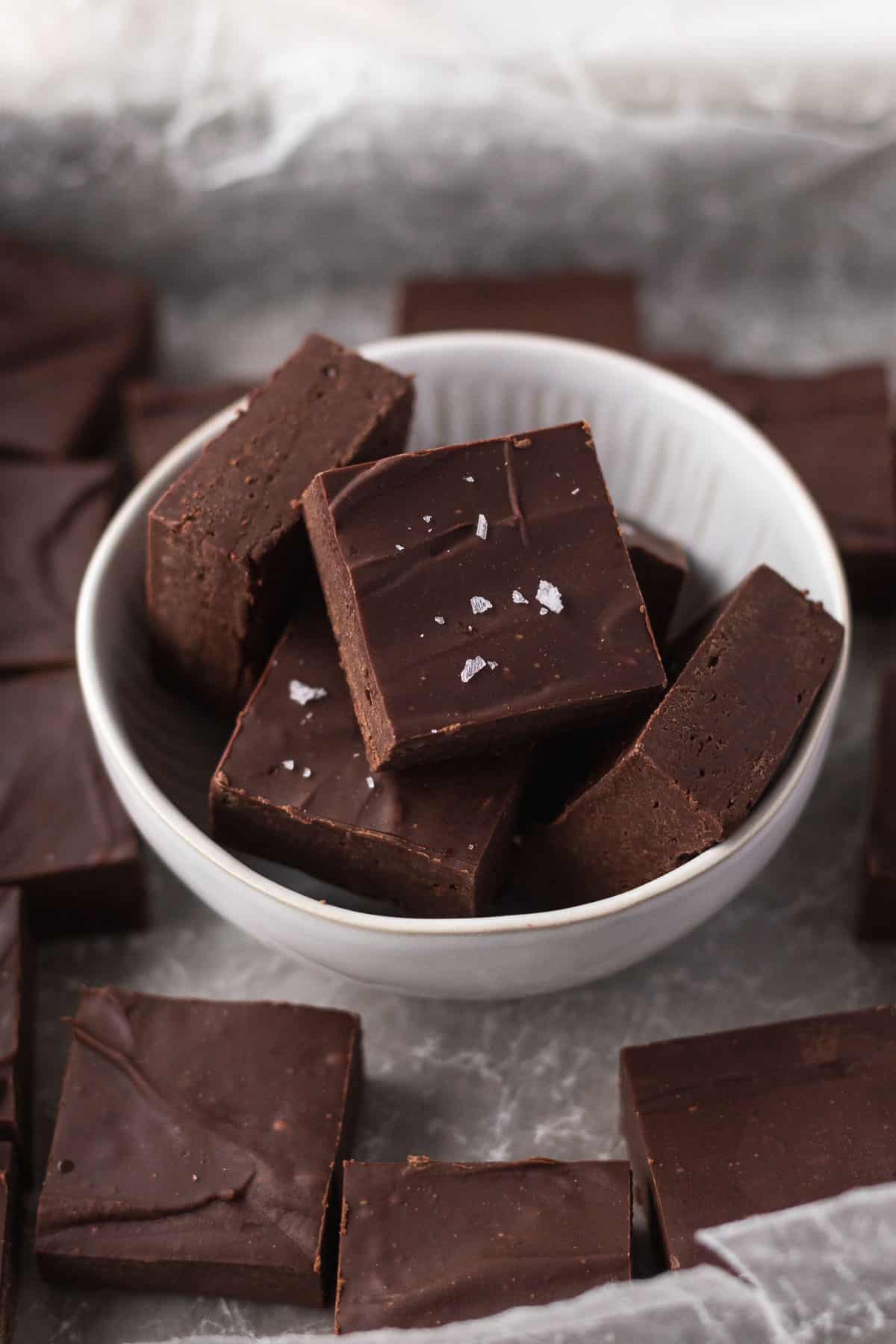 45 degree angle shot of cubes of dairy-free fudge in a white bowl on a piece of crumpled white parchment with other cubes of fudge.