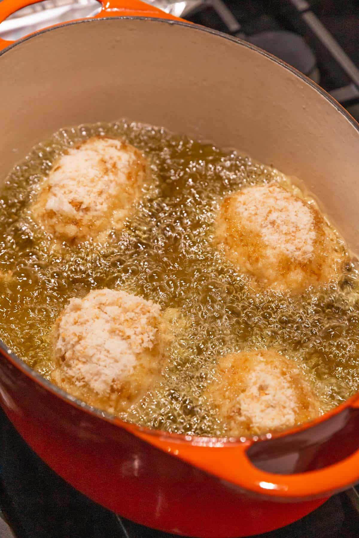 4 vegan potato croquettes frying in an orange enameled cast iron dutch oven. The bubbles are vigorous.