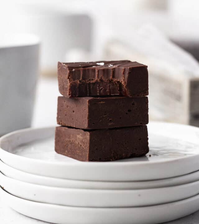 side on shot of 3 cubes of dairy-free fudge stacked on a stack of white plates with the top square having a bite taken out.