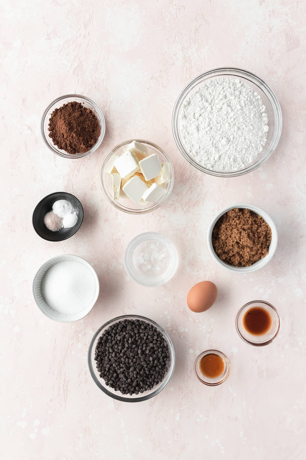 Ingredients to make double chocolate chip cookies measured out into small bowls and laid out on a pink surface.
