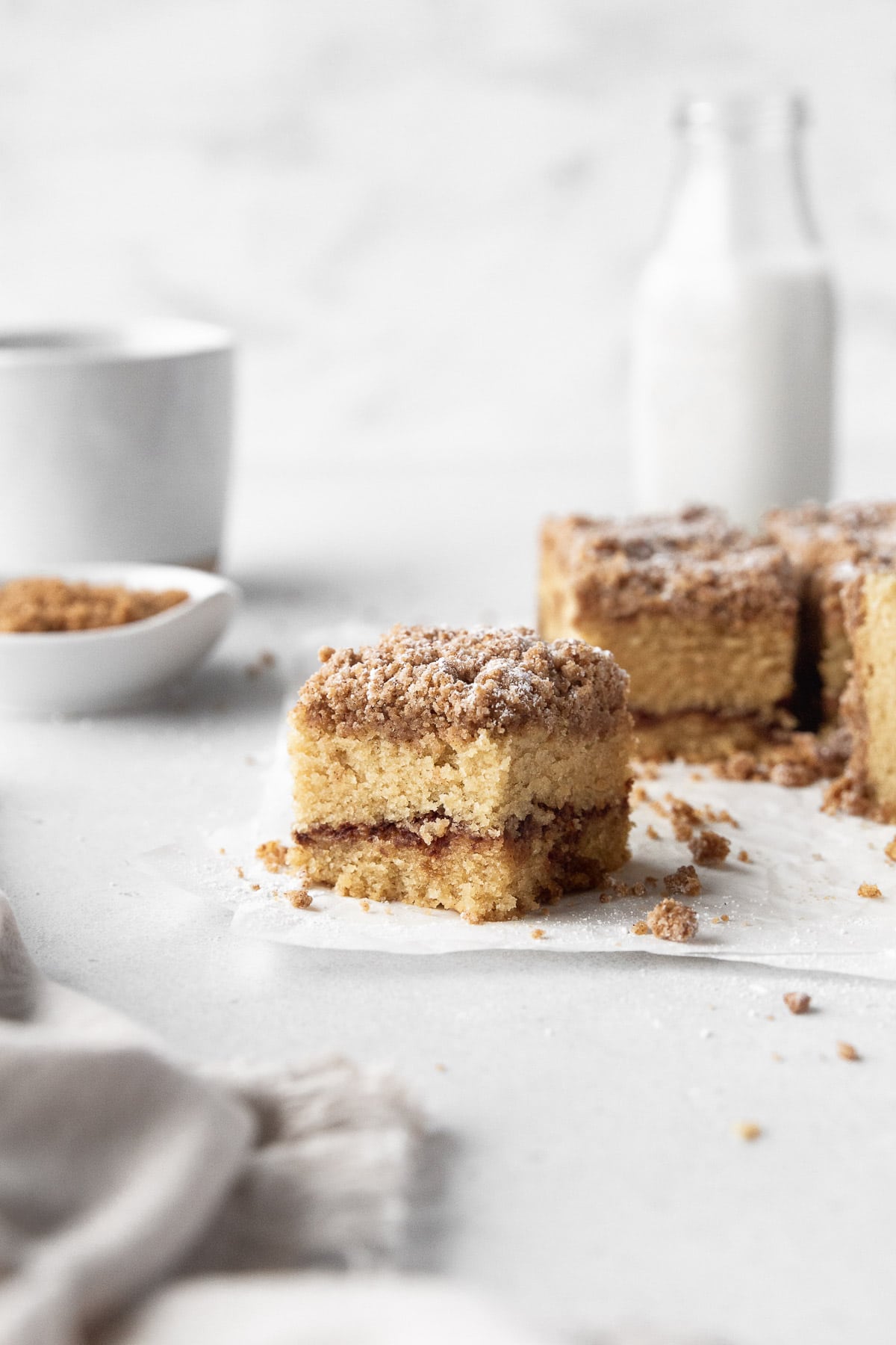 Square of GF cinnamon crumble cake with the remaining cake, extra crumble topping, and a jug of milk in the background.