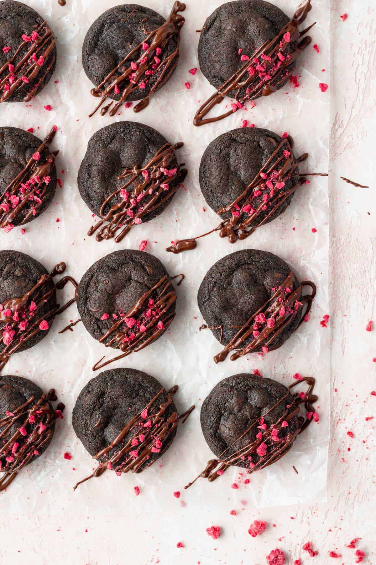 Chocolate chocolate cookies drizzled with chocolate and freeze-dried raspberries on top laid on a pink surface.