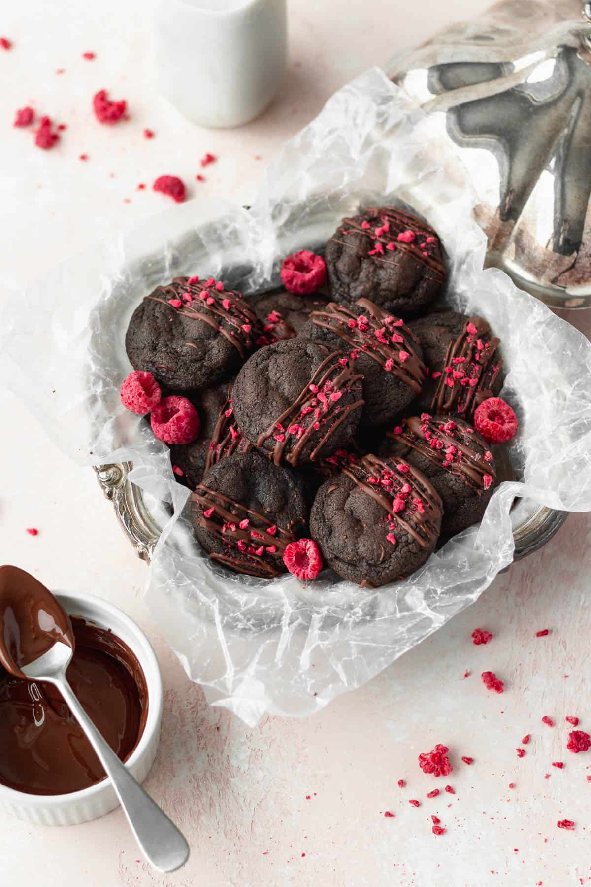 Overhead shot of double chocolate chip cookies in a silver dish lined with parchment paper and freeze-dried raspberries sprinkled all around.