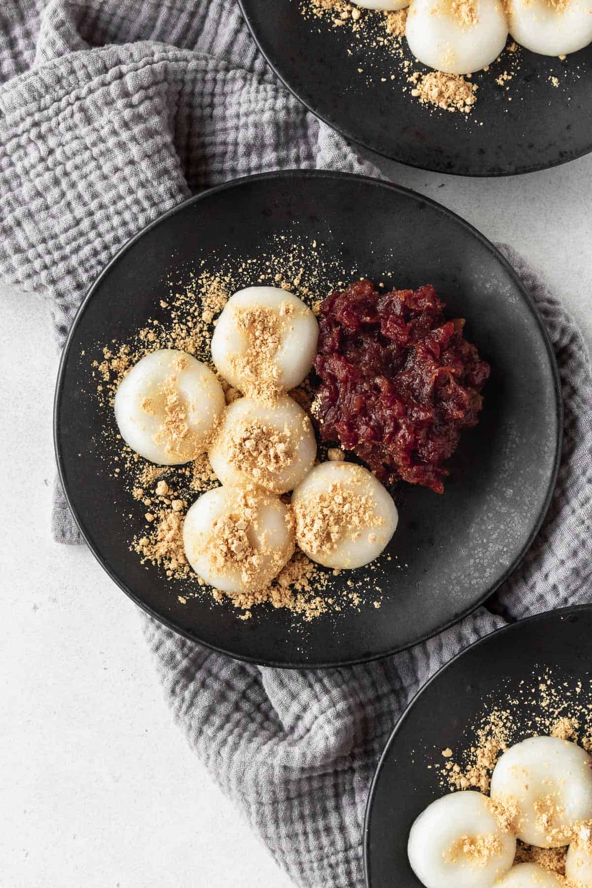An overhead shot of a black plate with shiratama dango, sweet red bean paste, and kinako sprinkled on top, with two more plates in the corner of the shot.