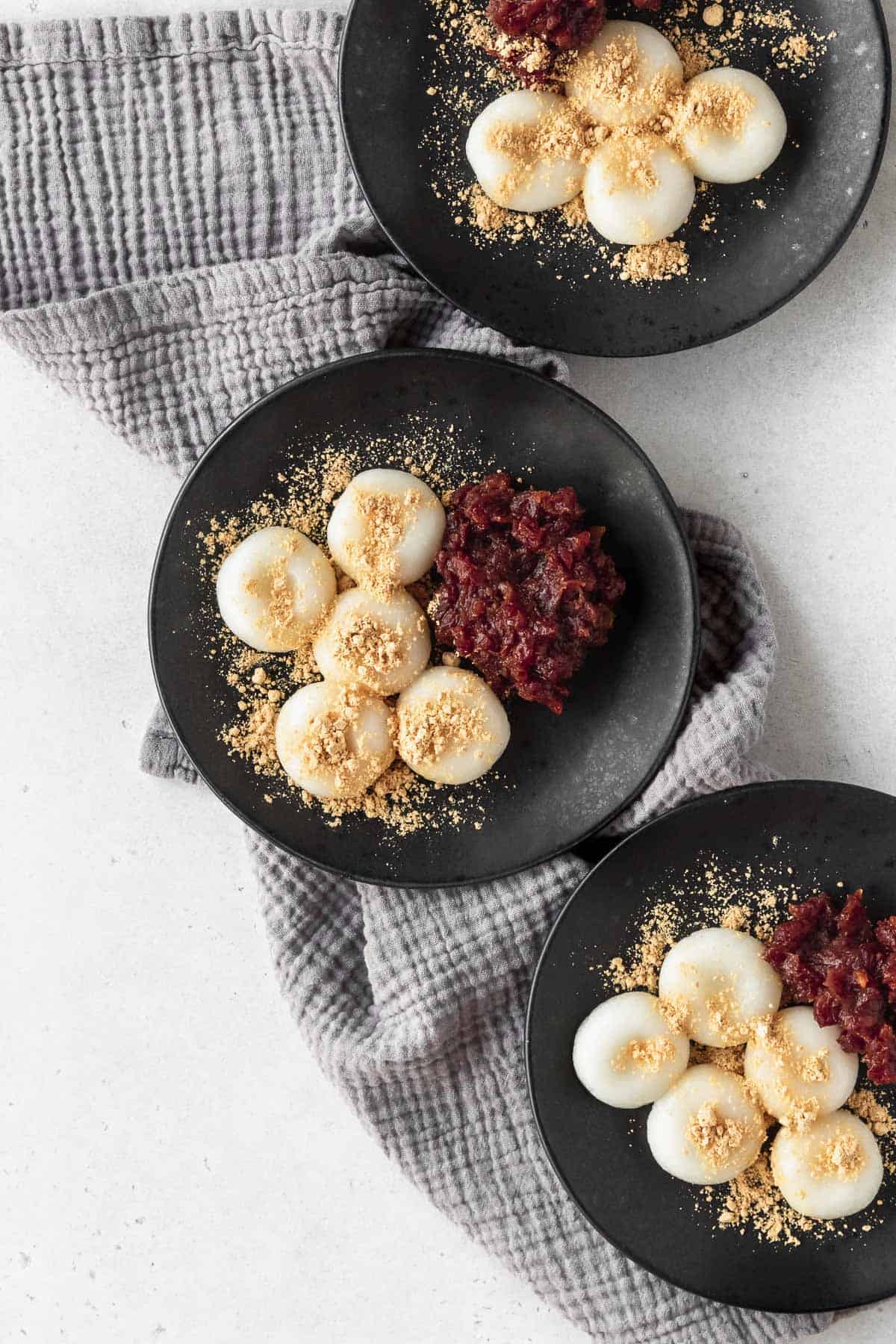 Shiratama on 3 black plates next to some sweet red bean paste and kinako sprinkled on top.