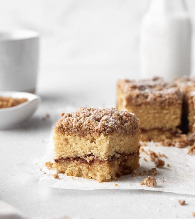 Square of GF cinnamon crumble cake with the remaining cake, extra crumble topping, and a jug of milk in the background.