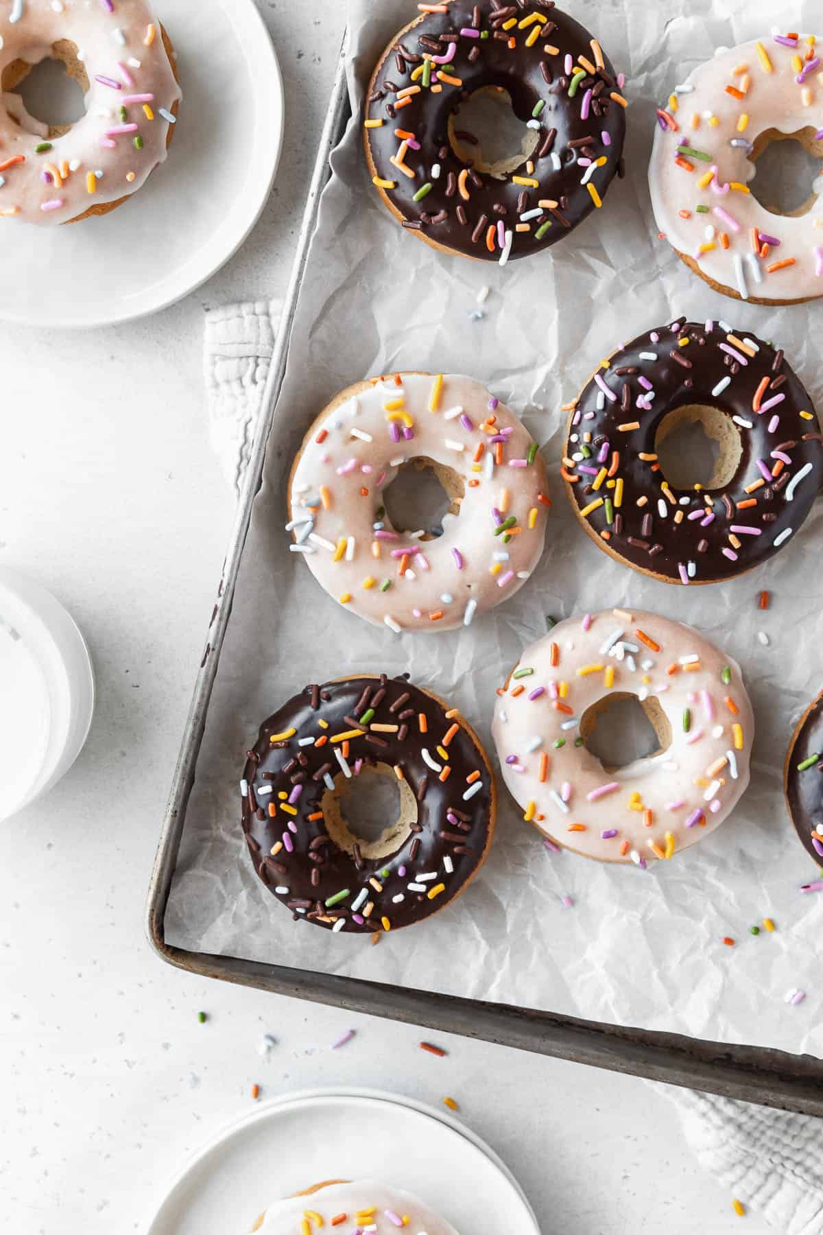 Overhead shot of a parchment lined tray of chocolate and vanilla dipped vegan baked donuts on a white table with small dessert plates holding vanilla glazed vegan doughnuts.