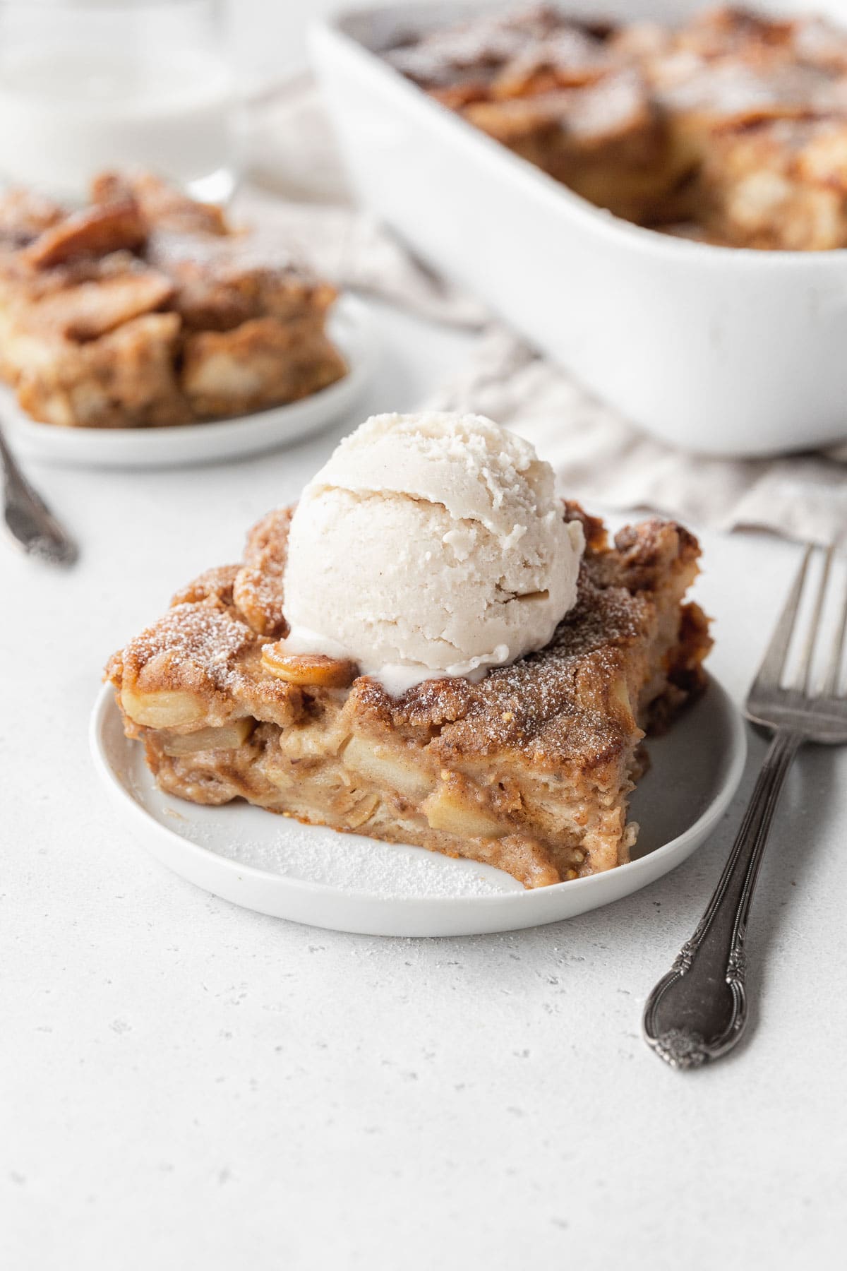 Slice of apple pie vegan bread pudding on a white dessert plate, topped with a scoop of vegan ice cream.