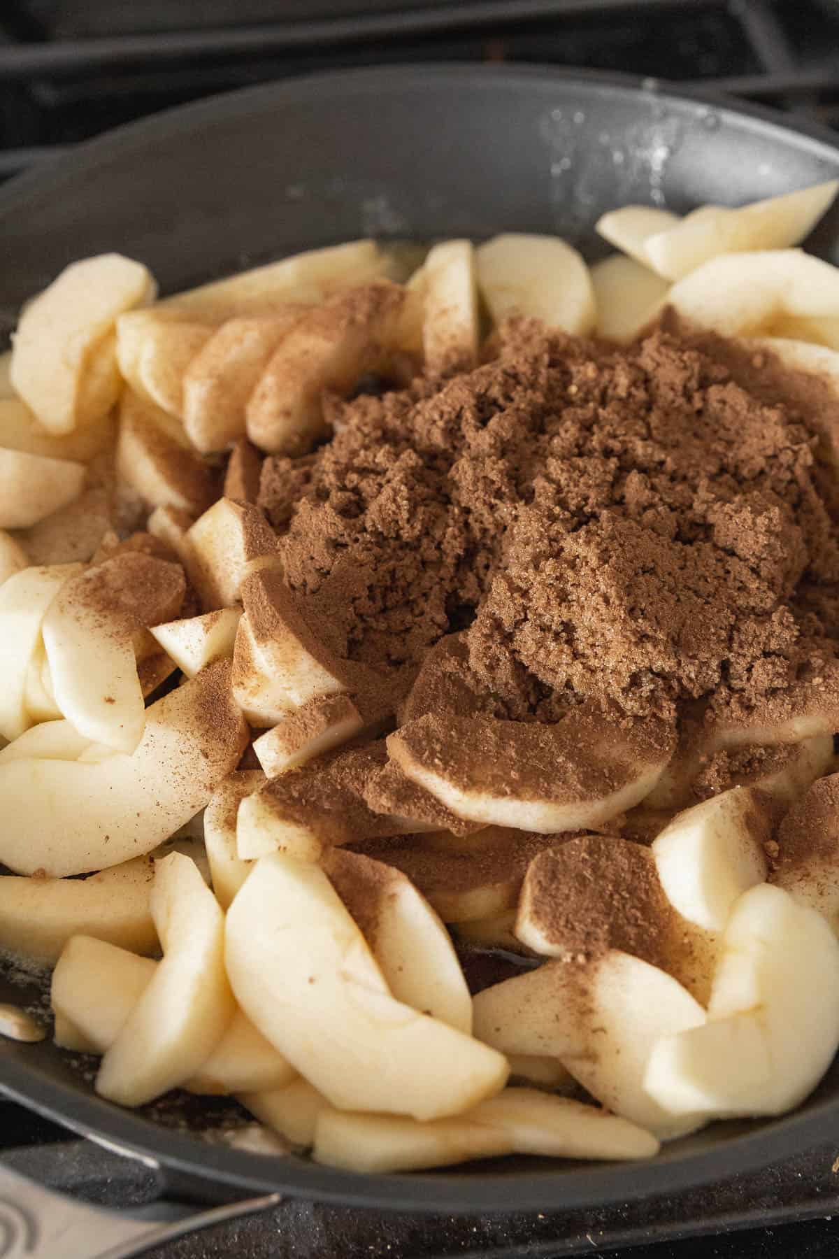 Slices of apples cooking with brown sugar and apple pie spice in a black pan.