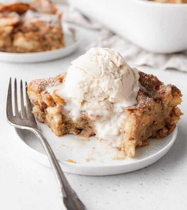 A partially eaten slice of vegan bread pudding on a white plate with a scoop of vanilla ice cream melting on top.