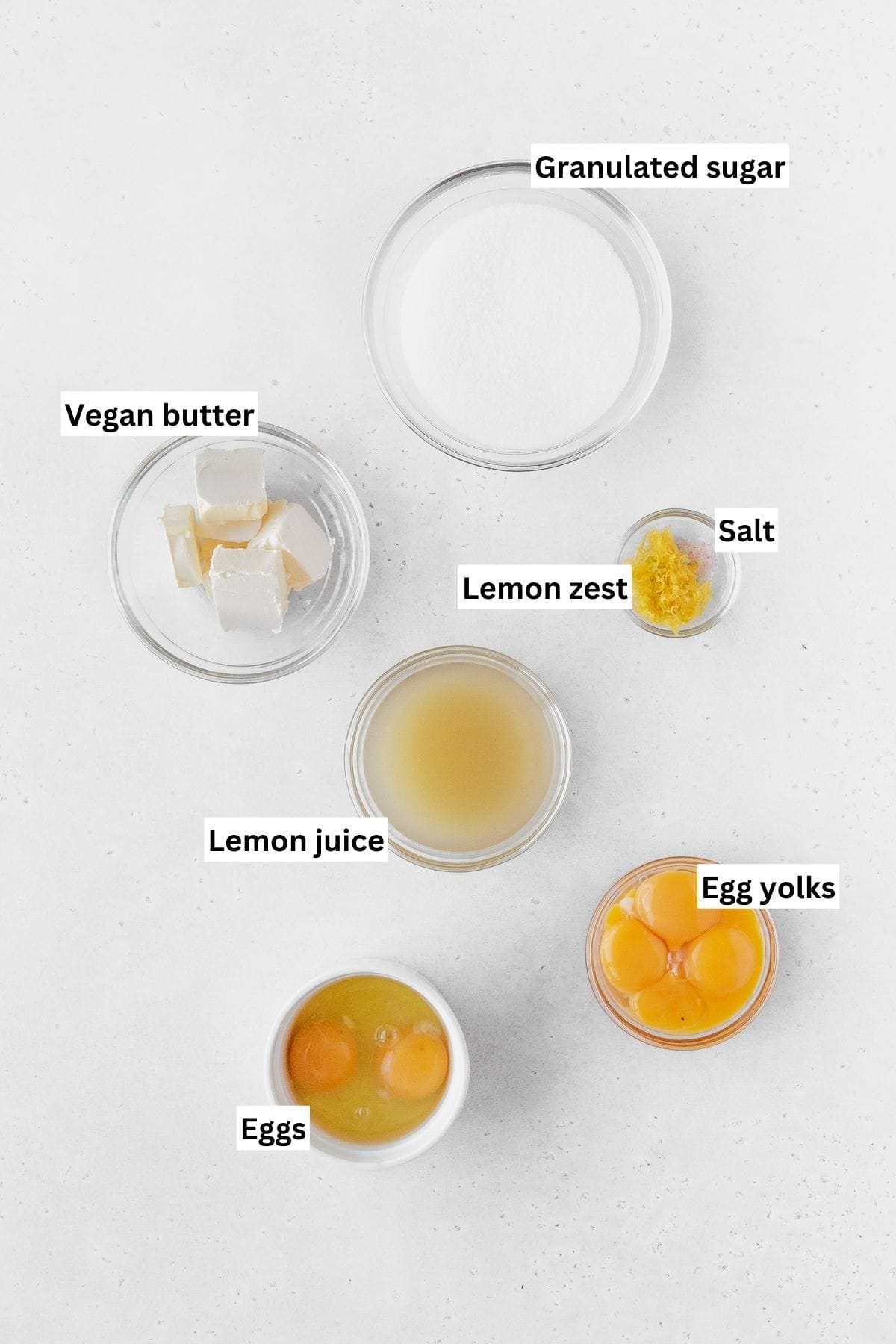 Lemon curd ingredients measured out into small glass bowls on a white table with text overlay.