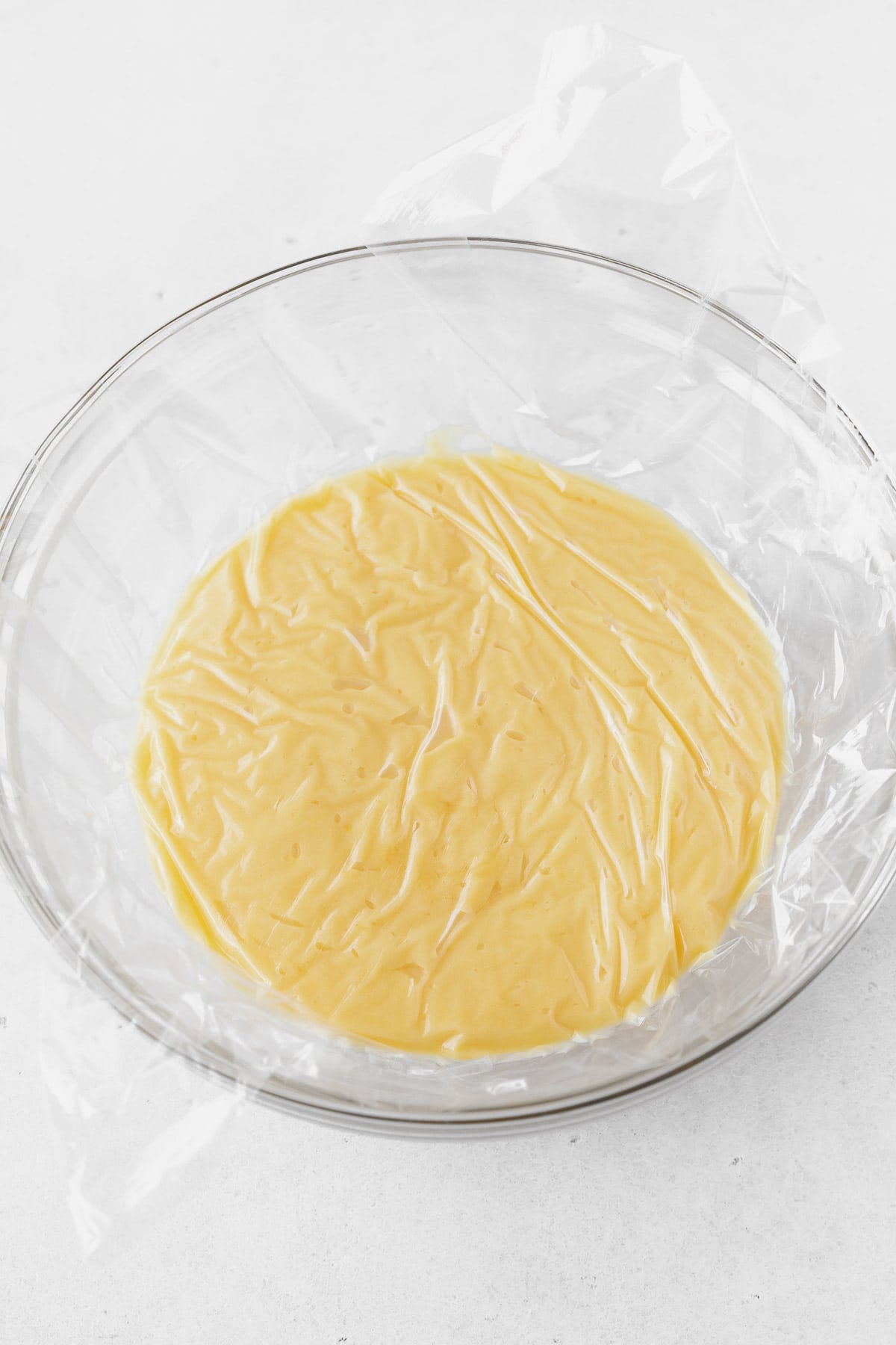 A glass bowl of lemon curd with a plastic wrap cover against the surface.