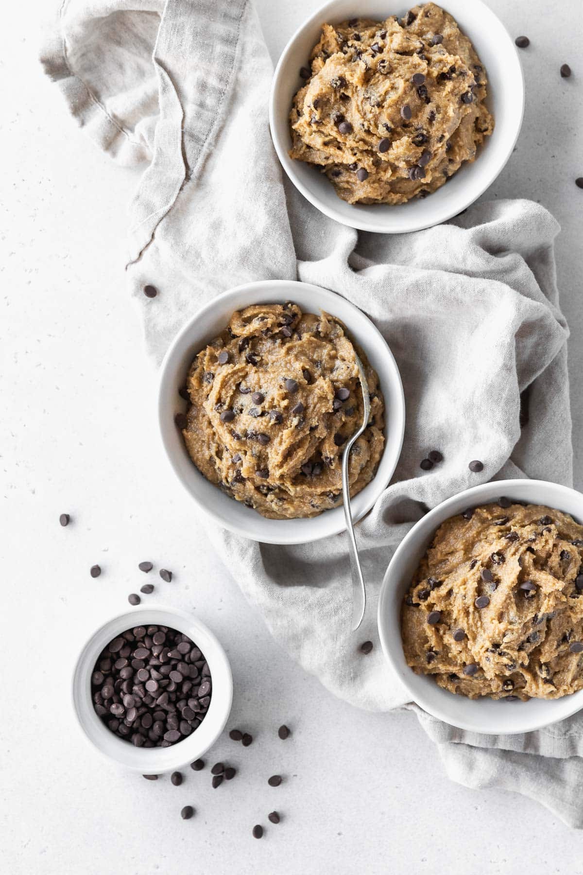 Overhead hero shot of 3 bowls of gluten-free cookie dough with silver spoon in the middle bowl and a small bowl of mini chocolate chips to the side.
