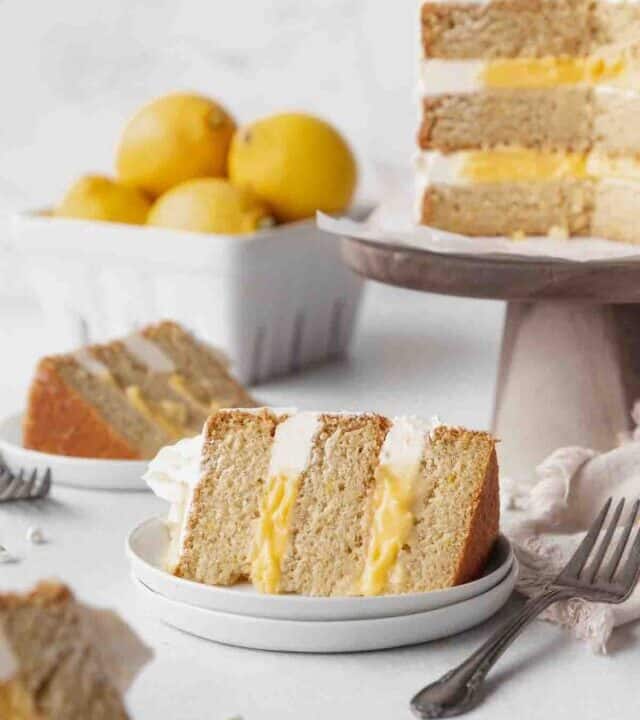 vertical hero shot of a slice of dairy-free lemon curd cake on a white dessert plate with a cake stand holding the rest of the cake and a ceramic fruit pint holding a handful of lemons in the background.