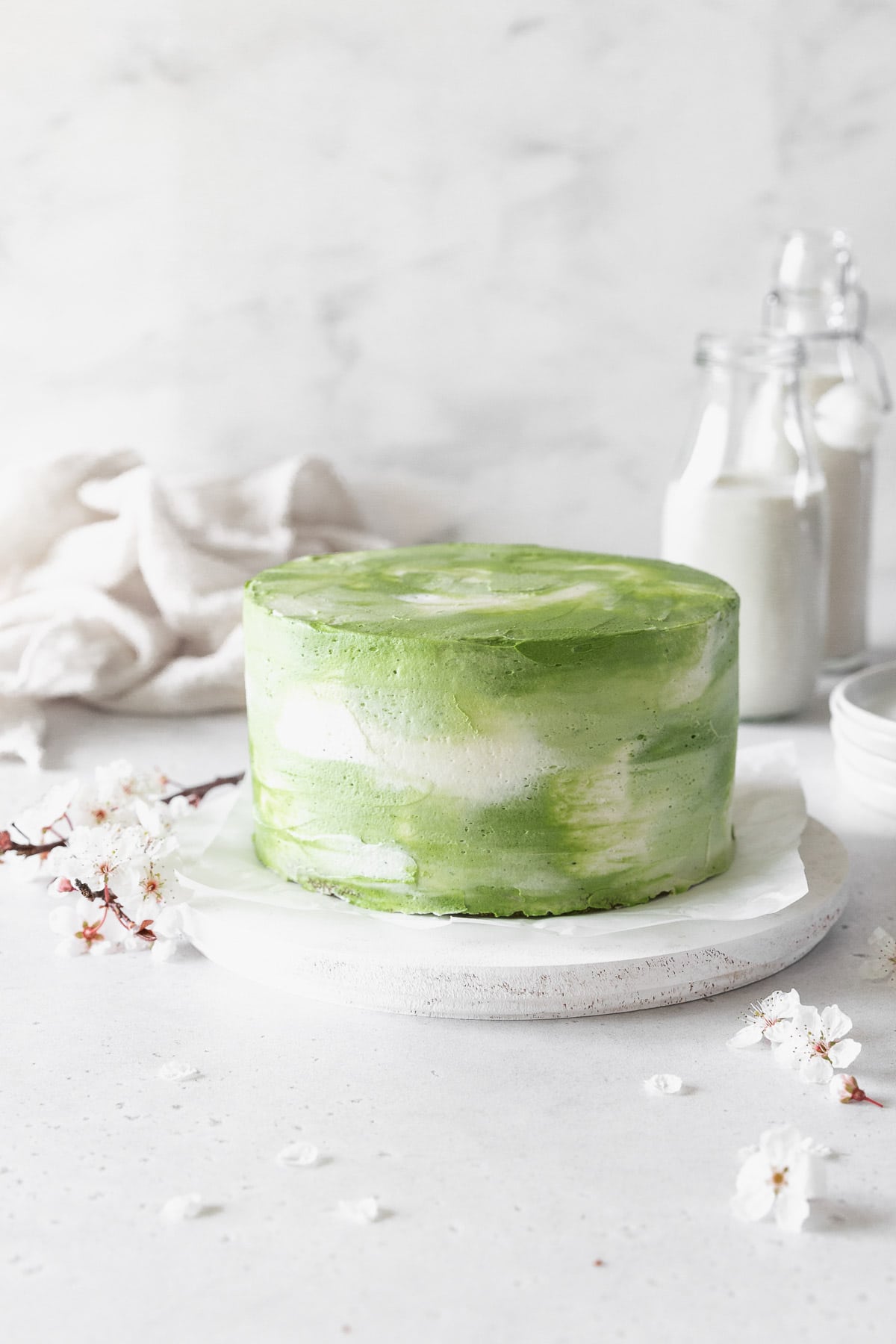 An iced matcha cake on a white cake stand with cherry blossoms. 