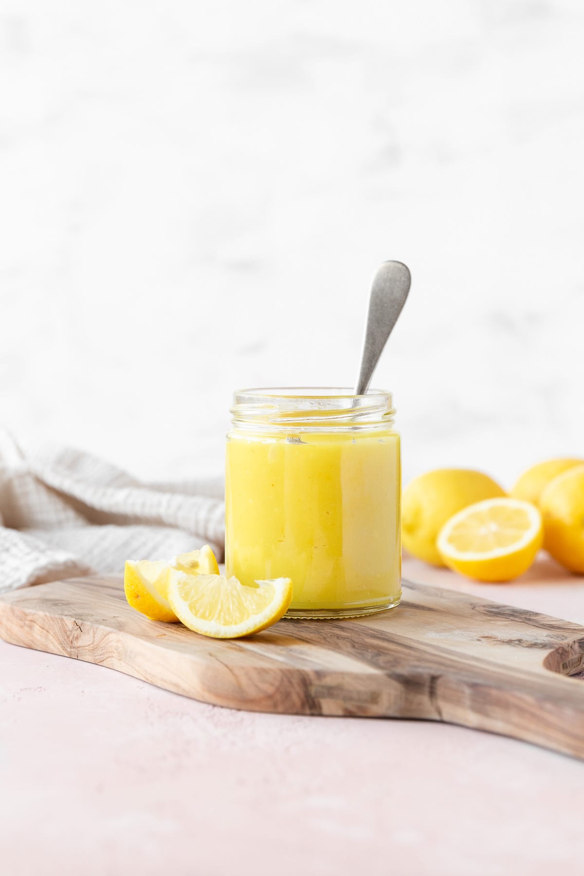 Side on shot of a small mason jar of vegan lemon curd with a silver spoon in it on a wooden cutting board with a few wedges of fresh lemon.