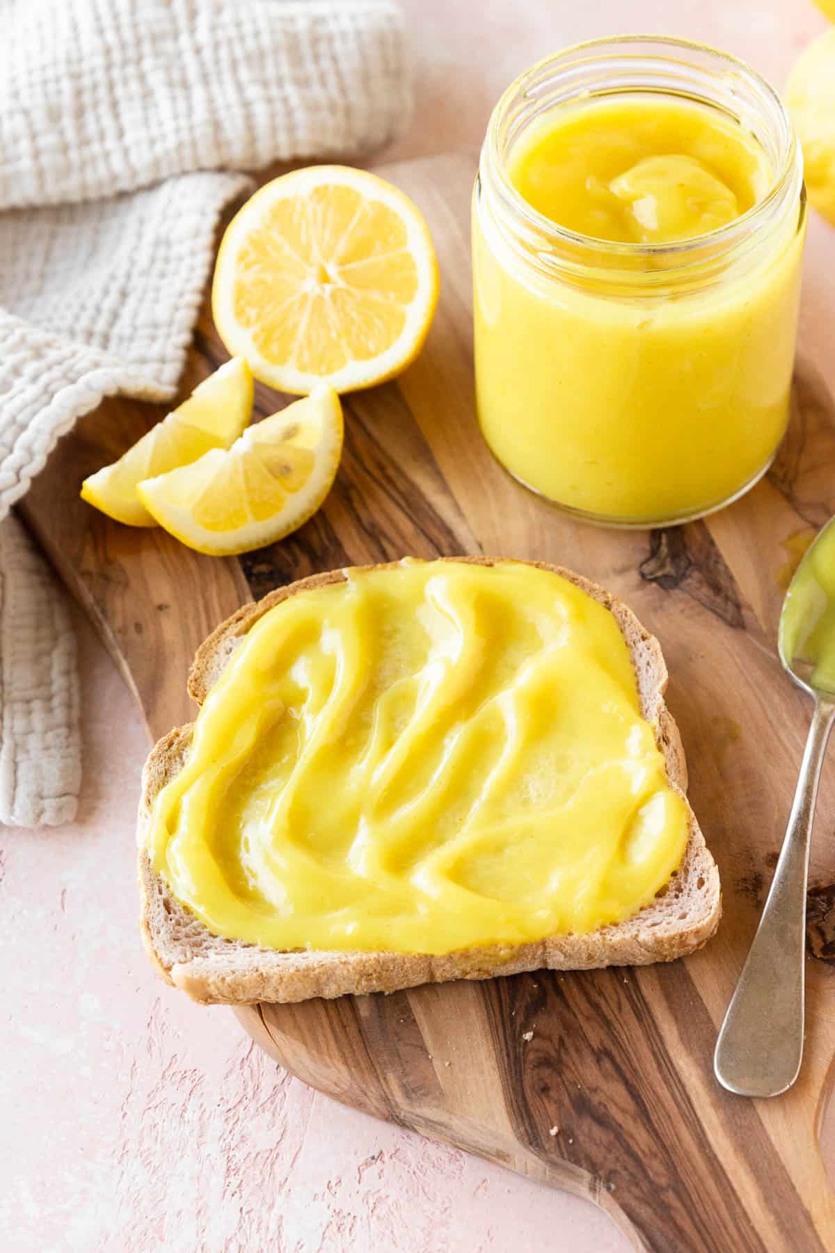 45 degree angle shot of a slice of toast spread with vegan lemon curd on a wooden cutting board with the jar of lemon curd and a halved lemon.