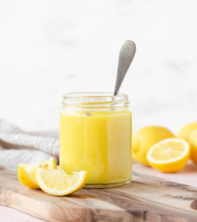 side on shot of a small mason jar of vegan lemon curd with a silver spoon in it on a wooden cutting board with a few wedges of fresh lemon.
