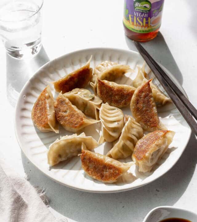 Angled shot of vegetable gyoza on a white plate with a glass of water and a bottle of vegan fish sauce in the background.