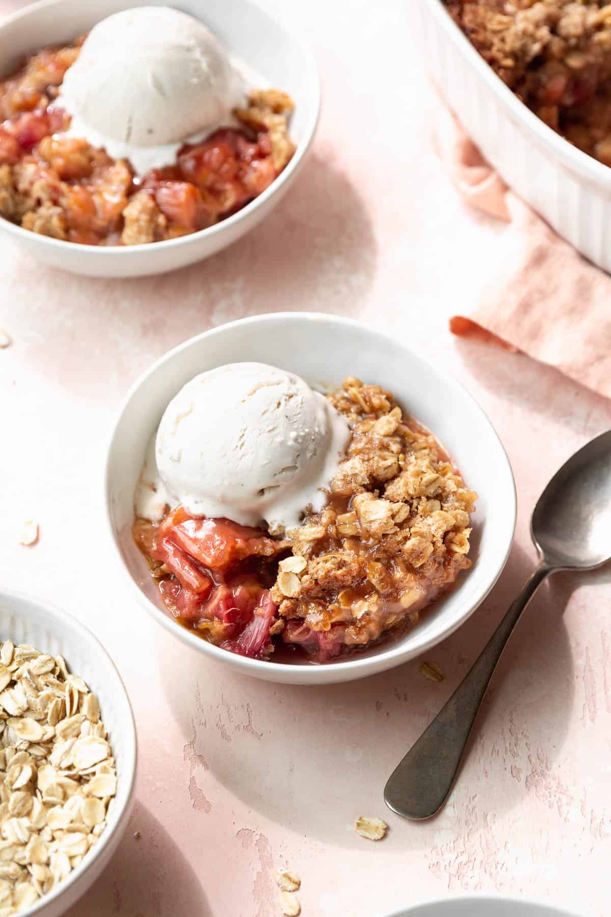 Two bowls of vegan, gluten-free rhubarb crisp with vanilla ice cream next to the larger casserole dish filled with the rest of the dessert.