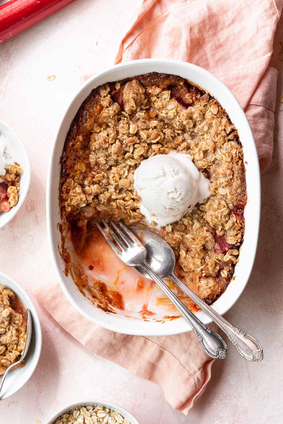 Overhead shot of a casserole dish filled with the baked GF rhubarb crisp with a few portions taken out and a silver fork and spoon in the gap.