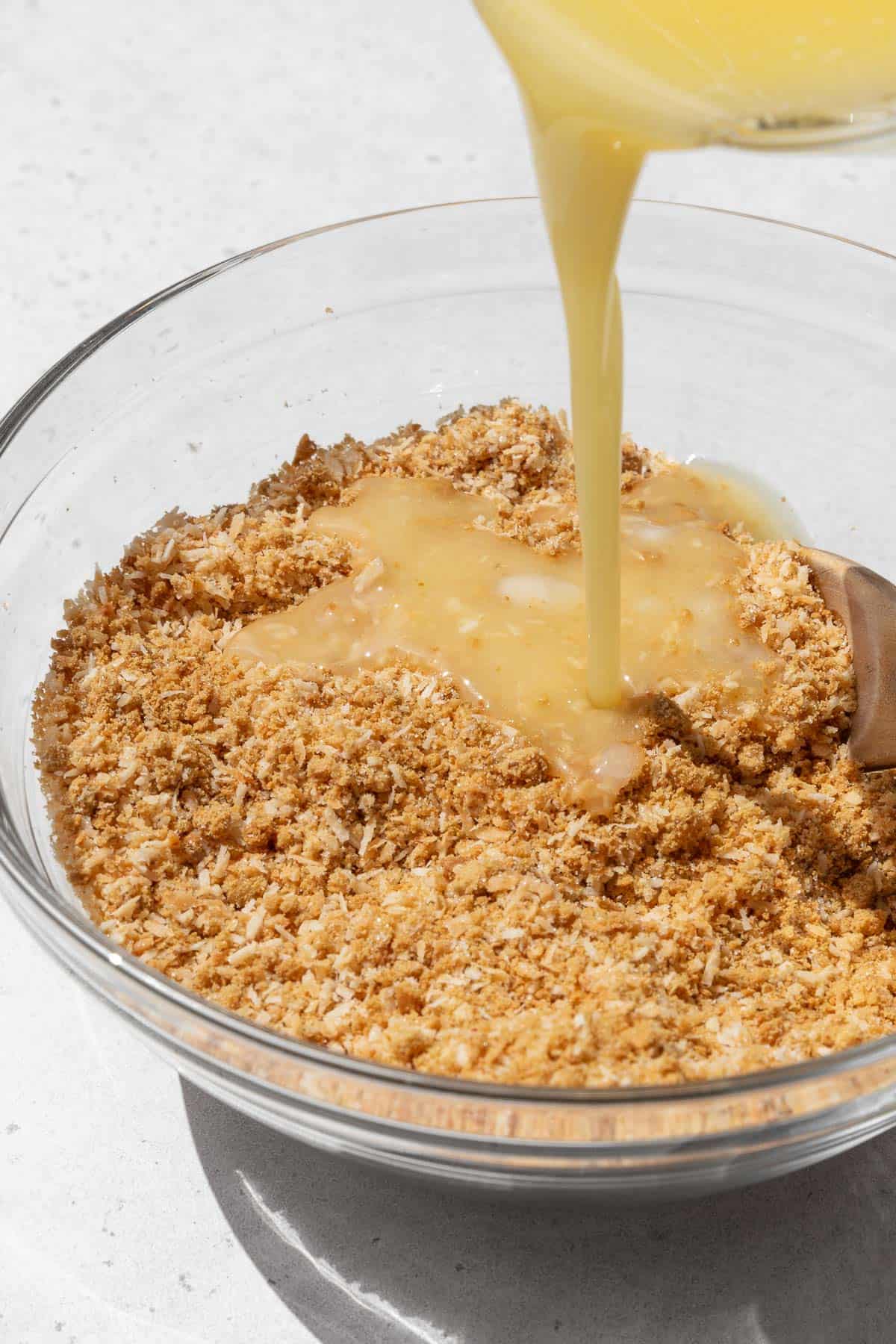 Action shot of melted vegan butter being poured into the bowl with the graham cracker crumb and toasted coconut mixture.