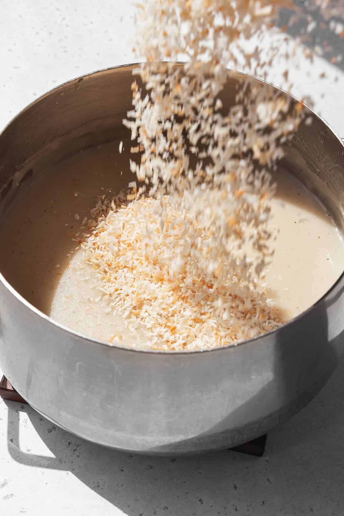 Action shot of toasted desiccated coconut being poured into the vegan custard base in the saucepan.