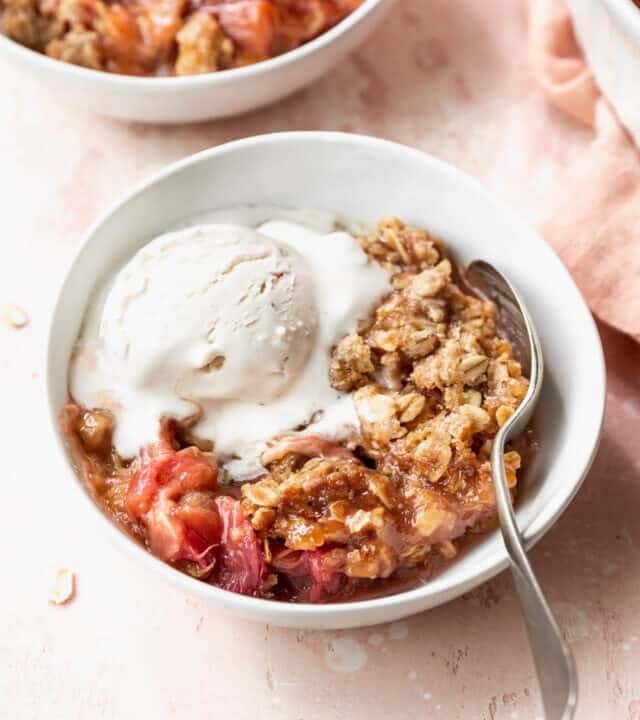 45 degree angle shot of a white bowl filled with gluten-free rhubarb crisp with oats crumble topping and a scoop of vanilla ice cream on a pink table.